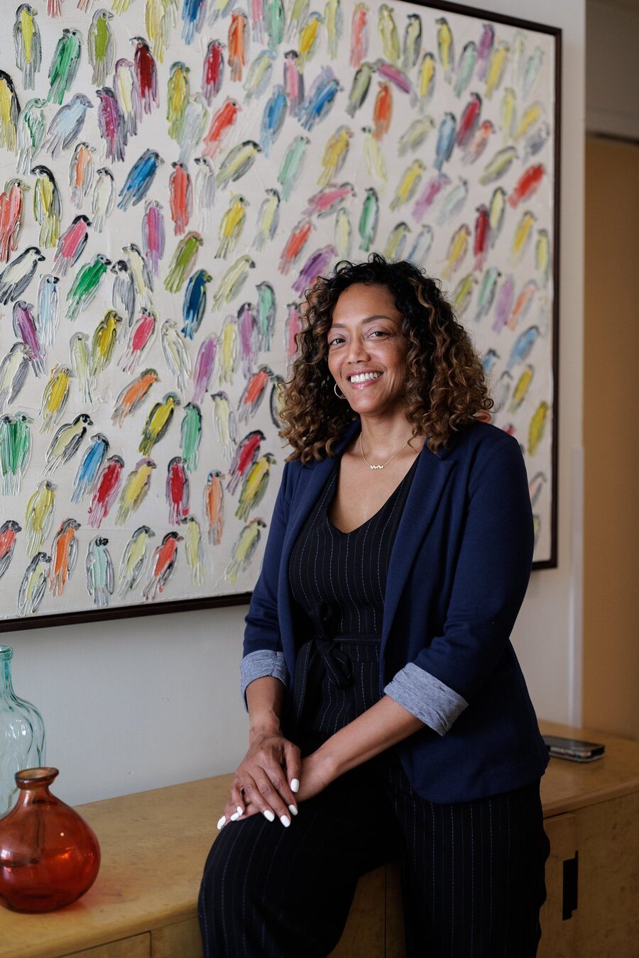 Akia Smith smiling, seated in front of a colorful painting of birds